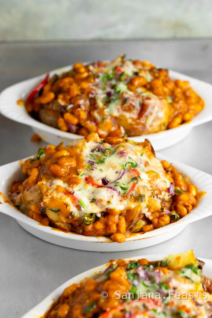 Jacket potatoes in a white dish topped with masala baked beans, melted cheese, chillies, onions and coriander.