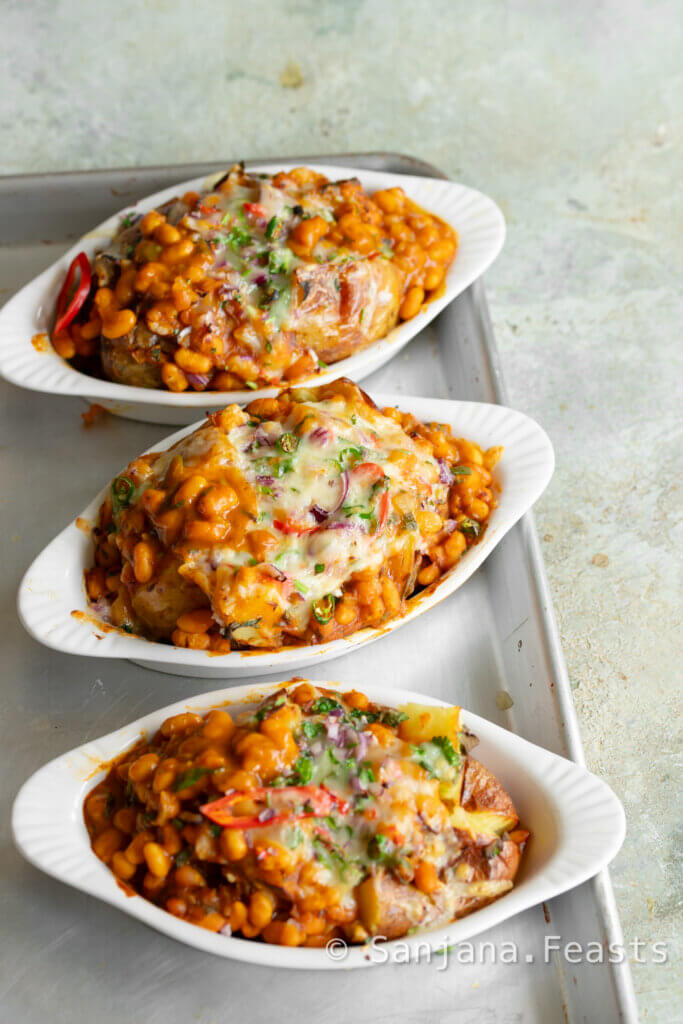 Jacket potatoes in three white gratin dishes topped with indian baked beans, melted cheese, chillies, onions and coriander.
