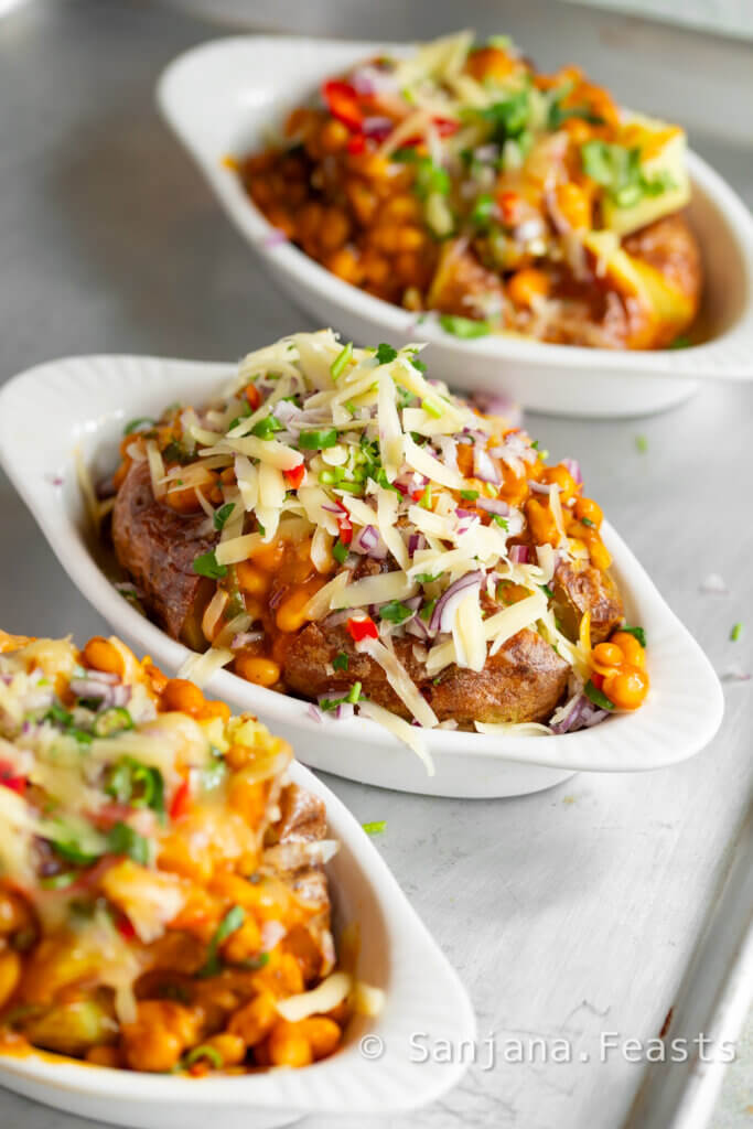 Jacket potato in a white dish topped with masala baked beans, cheddar cheese, chillies, onions and coriander.
